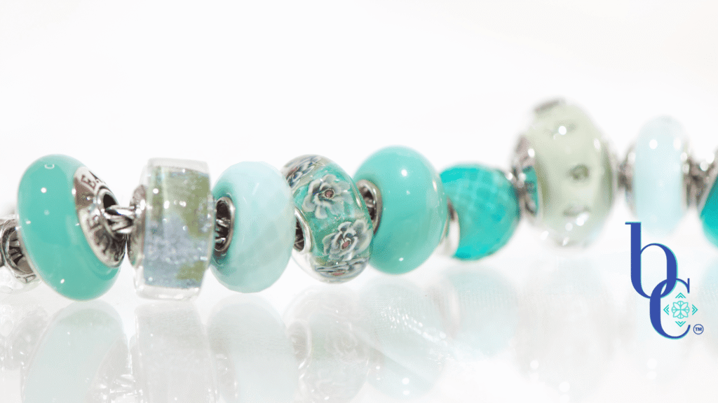 A macro photograph of a European charm bracelet resting on a highly reflective white surface. The stack features nine glass beads in saturated tones of sage green, icy mint, and vibrant teal, including a focal Elfbeads Azure glass bead. The background is a soft, luminous white with bokeh light circles.