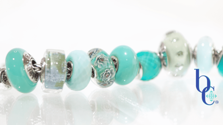 A macro photograph of a European charm bracelet resting on a highly reflective white surface. The stack features nine glass beads in saturated tones of sage green, icy mint, and vibrant teal, including a focal Elfbeads Azure glass bead. The background is a soft, luminous white with bokeh light circles.