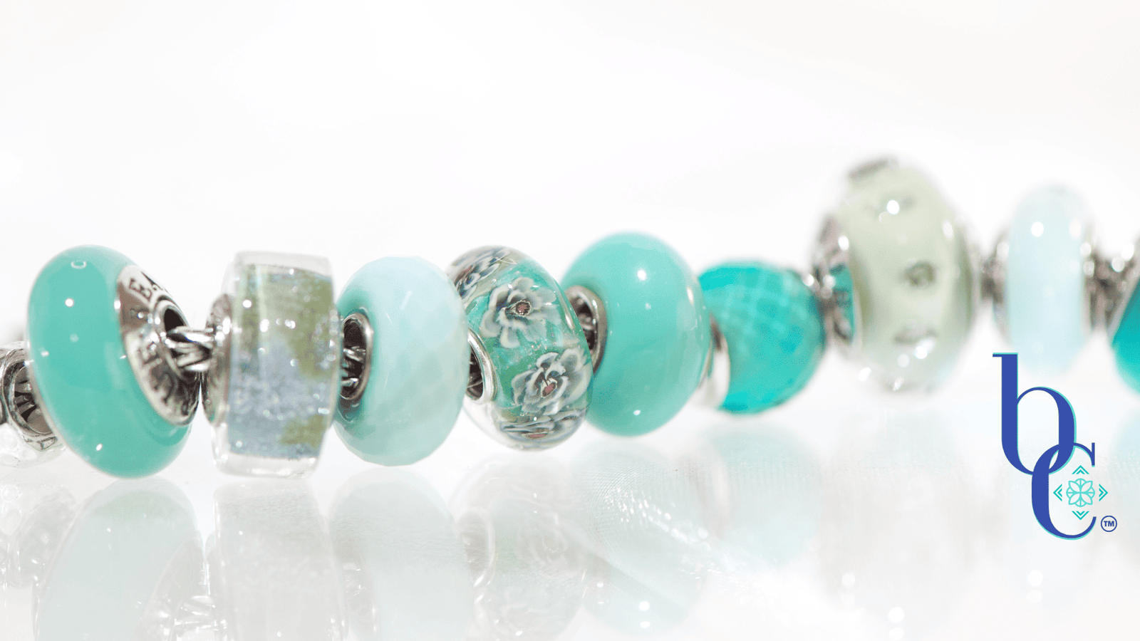 A macro photograph of a European charm bracelet resting on a highly reflective white surface. The stack features nine glass beads in saturated tones of sage green, icy mint, and vibrant teal, including a focal Elfbeads Azure glass bead. The background is a soft, luminous white with bokeh light circles.