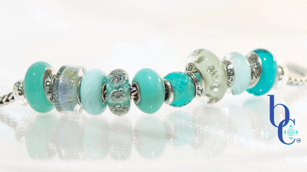  A macro photograph of a European charm bracelet resting on a highly reflective white surface. The stack features nine glass beads in saturated tones of sage green, icy mint, and vibrant teal, including a focal Elfbeads Azure glass bead. The background is a soft, luminous white with bokeh light circles.
