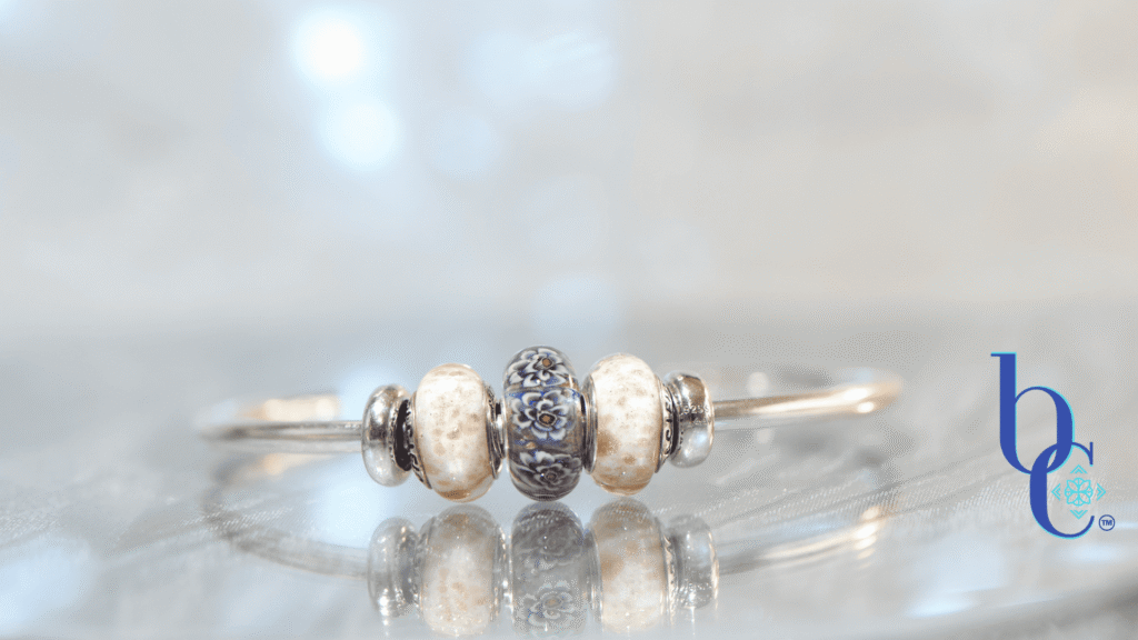 A macro studio photograph of an Elfbeads Rusty bead flanked by white and gold-flecked Pacific Brown Sugar beads on a silver bangle. The jewelry rests on a reflective dark grey surface with soft bokeh lighting in the background.