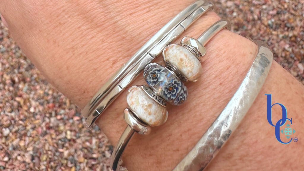A close-up of a woman's wrist wearing three silver bangles over wet, textured desert gravel. The center bangle features an Elfbeads Rusty glass charm—a dark grey floral design with a tiny orange center—flanked by two white and gold-flecked Pacific Brown Sugar glass beads.