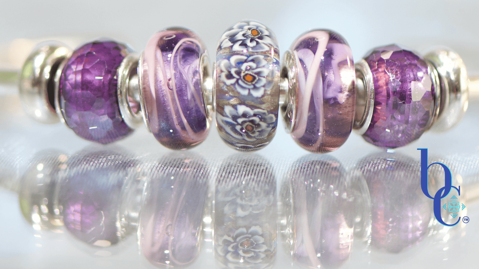 A macro studio photograph of an Elfbeads Rusty bead flanked by purple Chamilia Lilac Spin beads and faceted purple glass beads on a silver bangle. The jewelry rests on a reflective dark grey surface with soft bokeh lighting in the background.