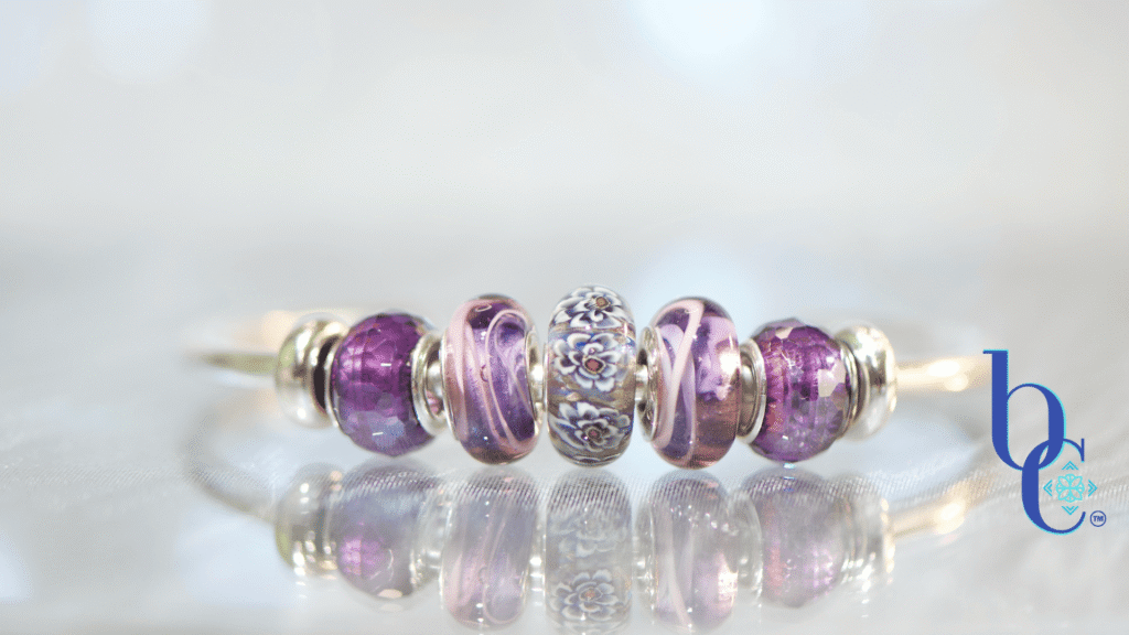 A macro studio photograph of an Elfbeads Rusty bead flanked by purple Chamilia Lilac Spin beads and faceted purple glass beads on a silver bangle. The jewelry rests on a reflective dark grey surface with soft bokeh lighting in the background.