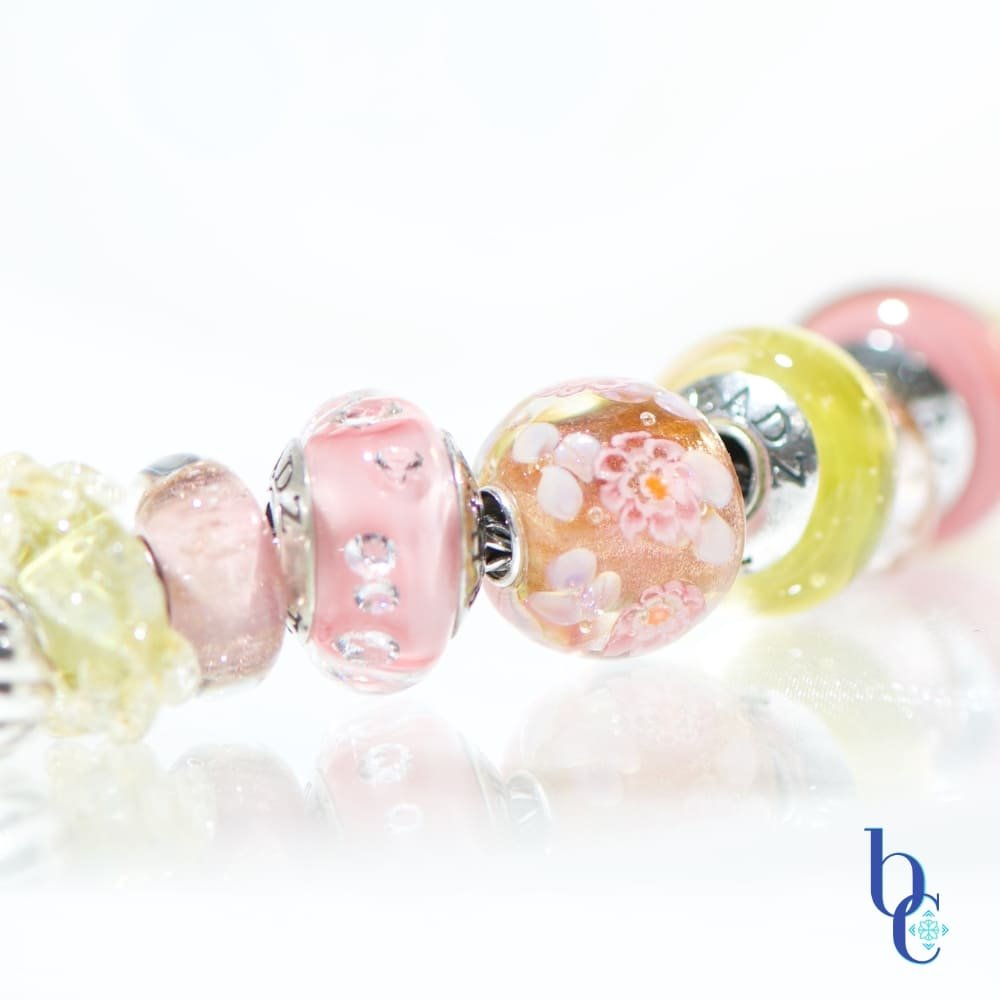 A macro photograph of the Amalfi Nectar European-style charm bracelet, featuring luminous yellow opal, soft mauve glass, and a central floral Elfbead resting on a bright, reflective white studio surface.