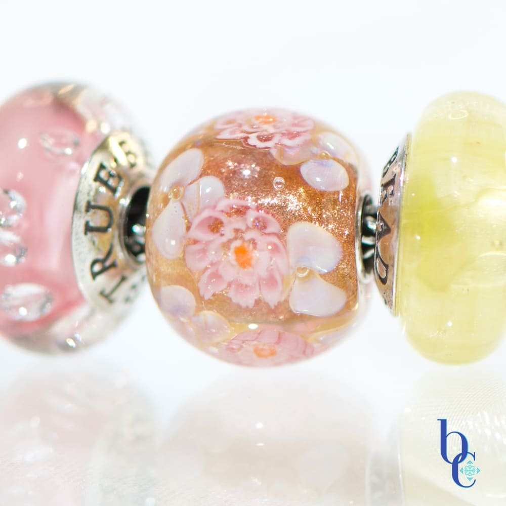 A macro photograph of the Amalfi Nectar European-style charm bracelet, featuring a focal of elfbeads golden sunset flowerworld glass resting on a bright, reflective white studio surface.