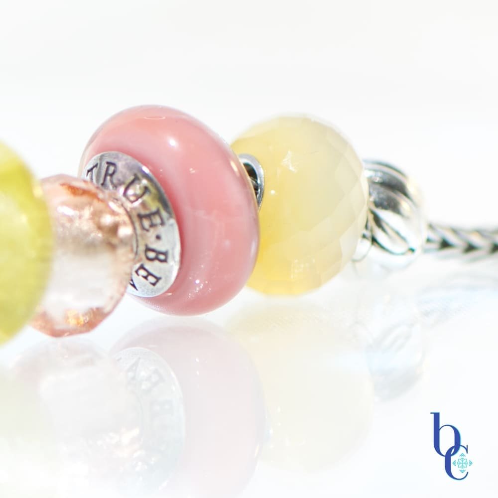 A macro photograph of the Amalfi Nectar European-style charm bracelet, featuring luminous soft mauve glass, and a faceted yellow chalcedony resting on a bright, reflective white studio surface.