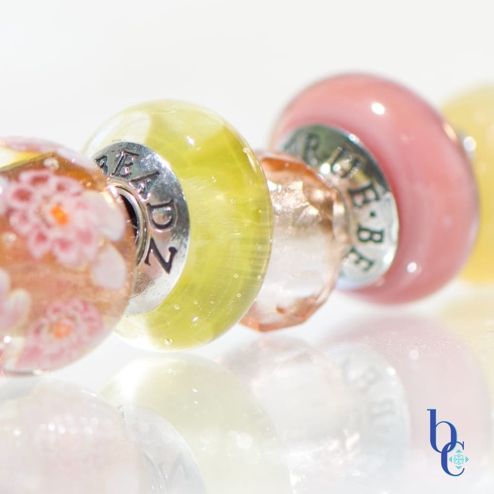 A macro photograph of the Amalfi Nectar European-style charm bracelet, featuring luminous soft mauve glass, and a pink prism resting on a bright, reflective white studio surface.