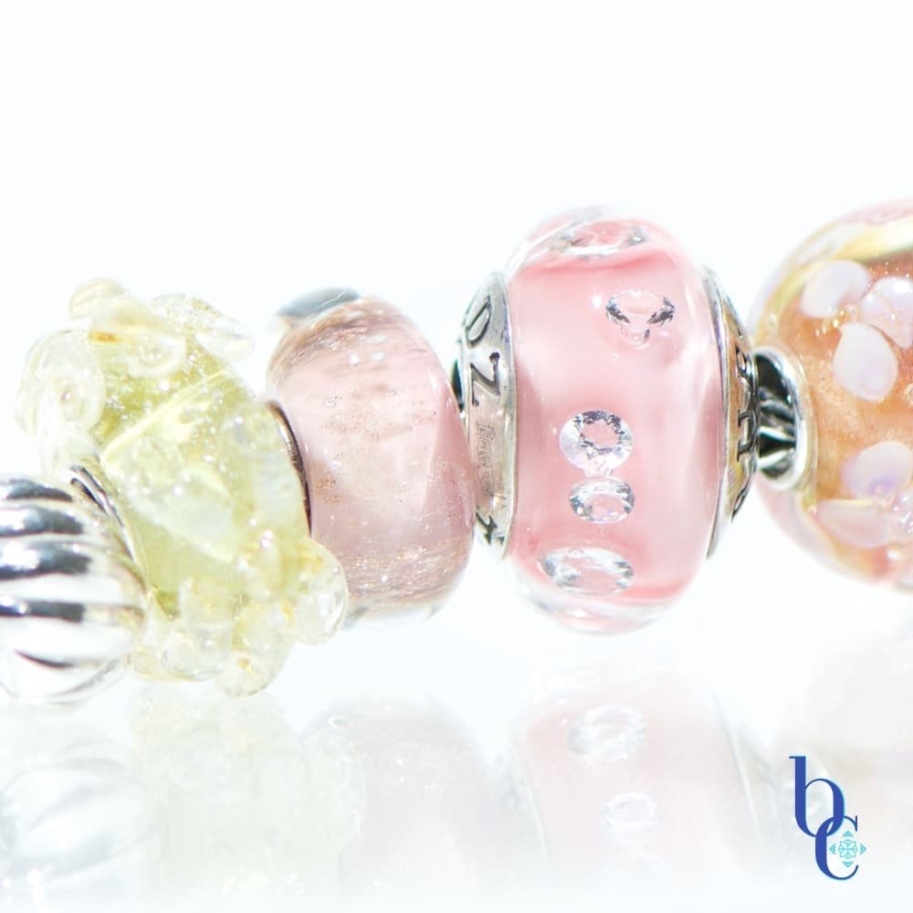 A macro photograph of the Amalfi Nectar European-style charm bracelet, featuring TrueBeadz pink CZ glass, and a Trollbeads pink desert resting on a bright, reflective white studio surface.
