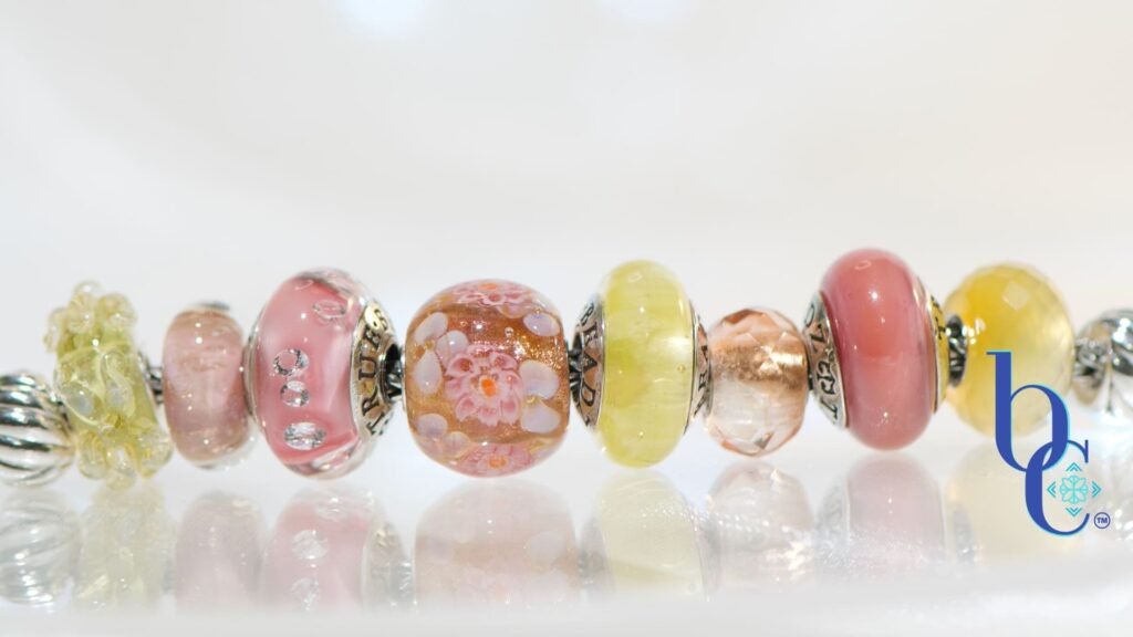 A macro photograph of the Amalfi Nectar European-style charm bracelet, featuring luminous yellow opal, soft mauve glass, and a central floral Elfbead resting on a bright, reflective white studio surface.