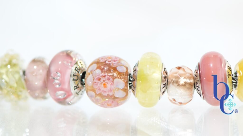 A macro photograph of the Amalfi Nectar European-style charm bracelet, featuring luminous yellow opal, soft mauve glass, and a central floral Elfbead resting on a bright, reflective white studio surface.