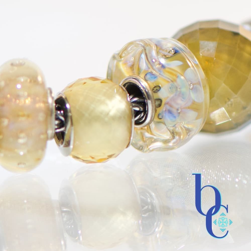 A macro studio photograph of a European-style charm bracelet resting on a highly reflective white surface, creating a clear mirror reflection below. The image showcases the beads on the center of the bracelet, highlighting their tactile textures and internal visual glow. The blue Bijoux Chat logo sits in the bottom right corner.
