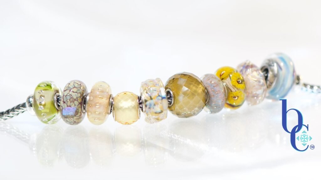 A macro studio photograph of a European-style charm bracelet resting on a highly reflective white surface, creating a clear mirror reflection below. The silver chain holds ten glass beads in dried botanical tones of translucent goldenrod, dusty plum, and faceted warm olive, highlighting their tactile textures and internal visual glow. The blue Bijoux Chat logo sits in the bottom right corner.