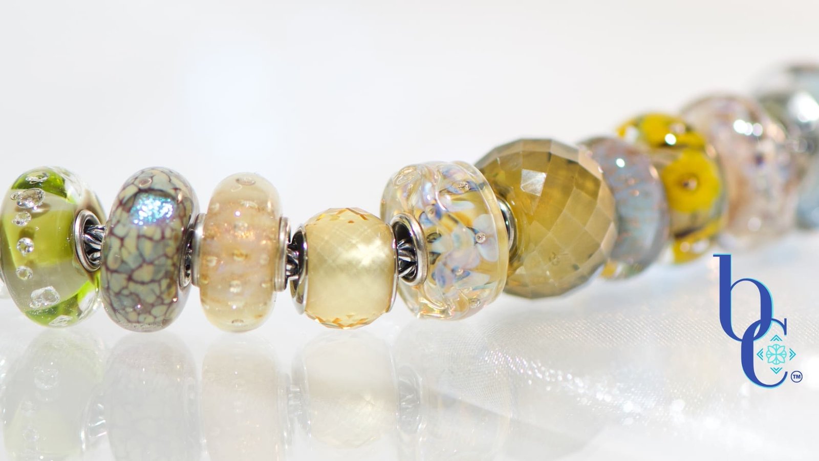 A macro studio photograph of a European-style charm bracelet resting on a highly reflective white surface, creating a clear mirror reflection below. The silver chain holds ten glass beads in dried botanical tones of translucent goldenrod, dusty plum, and faceted warm olive, highlighting their tactile textures and internal visual glow. The blue Bijoux Chat logo sits in the bottom right corner.