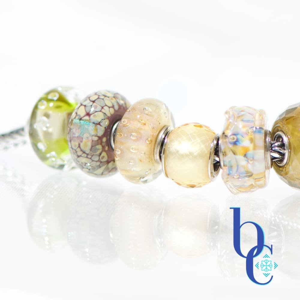 A macro studio photograph of a European-style charm bracelet resting on a highly reflective white surface, creating a clear mirror reflection below. The image showcases the beads on the left side of the bracelet, highlighting their tactile textures and internal visual glow. The blue Bijoux Chat logo sits in the bottom right corner.