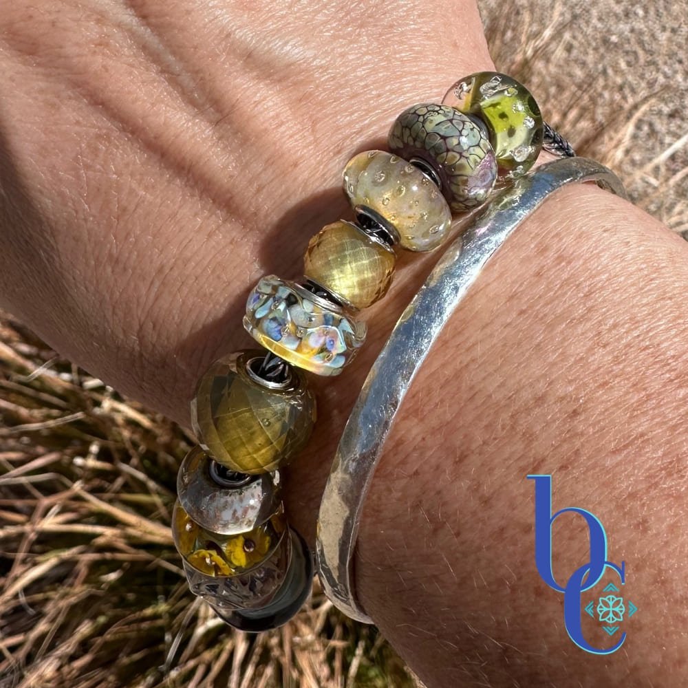 A macro lifestyle photograph of a European-style charm bracelet resting on a woman's wrist outdoors in the bright sunlight. The silver chain holds ten glass beads in dried botanical tones of translucent goldenrod, dusty plum, and faceted warm olive, highlighting their tactile textures and internal visual glow. The blue Bijoux Chat logo sits in the bottom right corner.