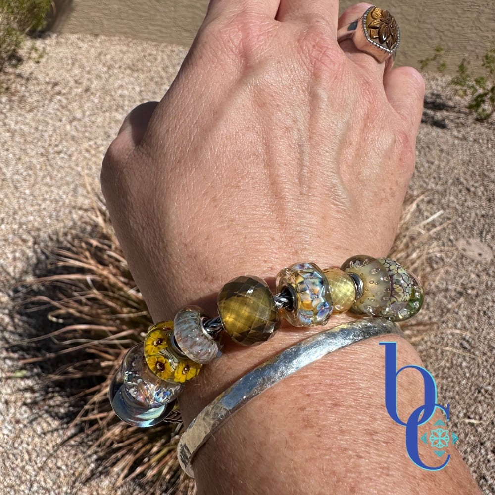 A macro lifestyle photograph of a European-style charm bracelet resting on a woman's wrist outdoors in the bright sunlight. The silver chain holds ten glass beads in dried botanical tones of translucent goldenrod, dusty plum, and faceted warm olive, highlighting their tactile textures and internal visual glow. The blue Bijoux Chat logo sits in the bottom right corner.