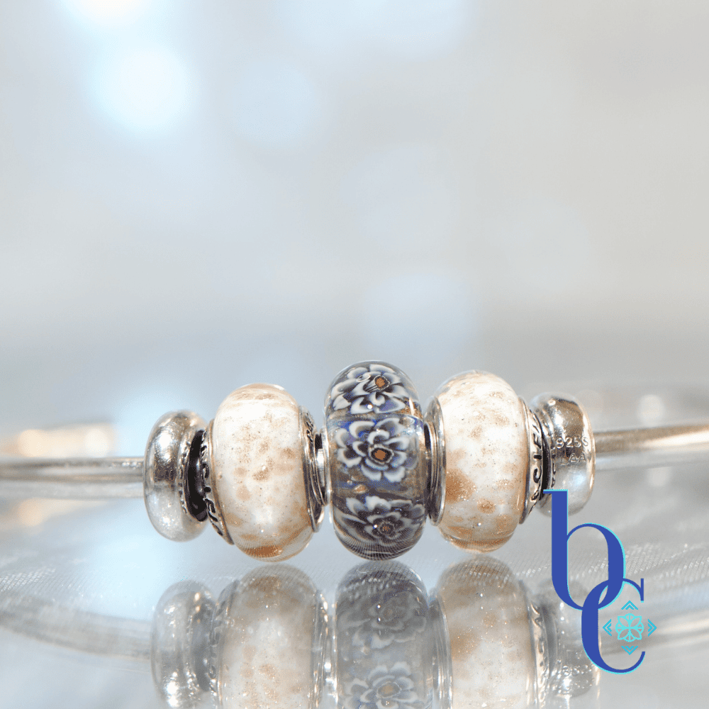 A macro studio photograph of an Elfbeads Rusty bead flanked by white and gold-flecked Pacific Brown Sugar beads on a silver bangle. The jewelry rests on a reflective dark grey surface with soft bokeh lighting in the background.