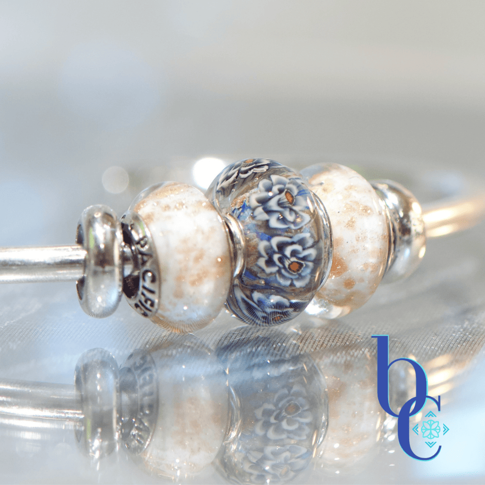 A macro studio photograph of an Elfbeads Rusty bead flanked by white and gold-flecked Pacific Brown Sugar beads on a silver bangle. The jewelry rests on a reflective dark grey surface with soft bokeh lighting in the background.