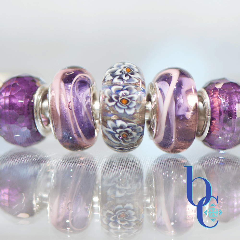 A macro studio photograph of an Elfbeads Rusty bead flanked by purple Chamilia Lilac Spin beads and faceted purple glass beads on a silver bangle. The jewelry rests on a reflective dark grey surface with soft bokeh lighting in the background.