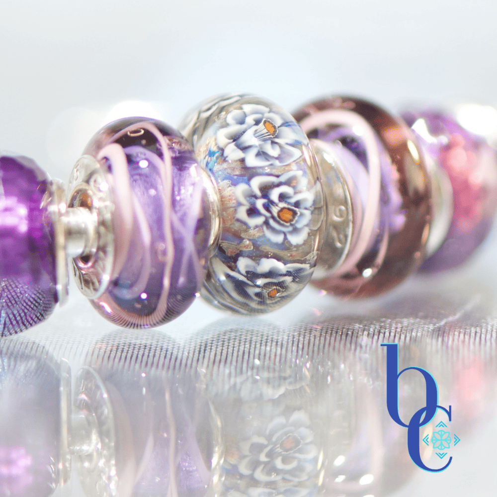 A macro studio photograph of an Elfbeads Rusty bead flanked by purple Chamilia Lilac Spin beads and faceted purple glass beads on a silver bangle. The jewelry rests on a reflective dark grey surface with soft bokeh lighting in the background.