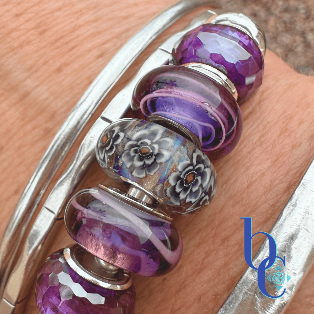 A close-up of a woman's wrist wearing three silver bangles over wet desert gravel. The center bangle features a dark grey and floral Elfbeads Rusty glass charm, surrounded by bright purple Chamilia Lilac Spin beads and deep purple faceted glass beads.