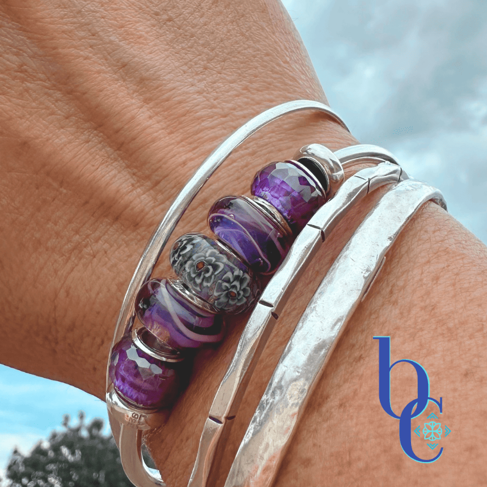 A close-up of a woman's wrist wearing three silver bangles over wet desert gravel. The center bangle features a dark grey and floral Elfbeads Rusty glass charm, surrounded by bright purple Chamilia Lilac Spin beads and deep purple faceted glass beads.