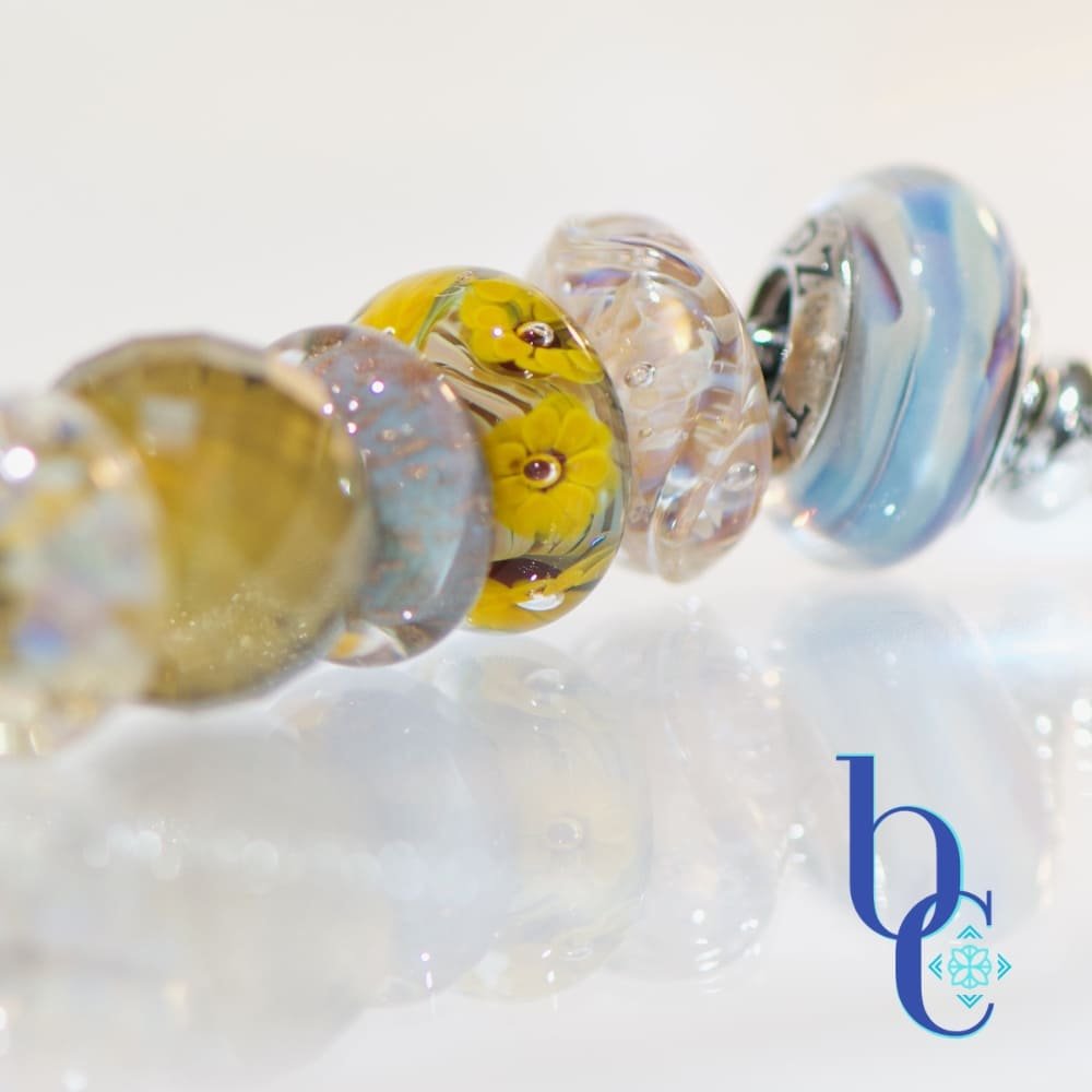 A macro studio photograph of a European-style charm bracelet resting on a highly reflective white surface, creating a clear mirror reflection below. The image showcases the beads on the right side of the bracelet, highlighting their tactile textures and internal visual glow. The blue Bijoux Chat logo sits in the bottom right corner.