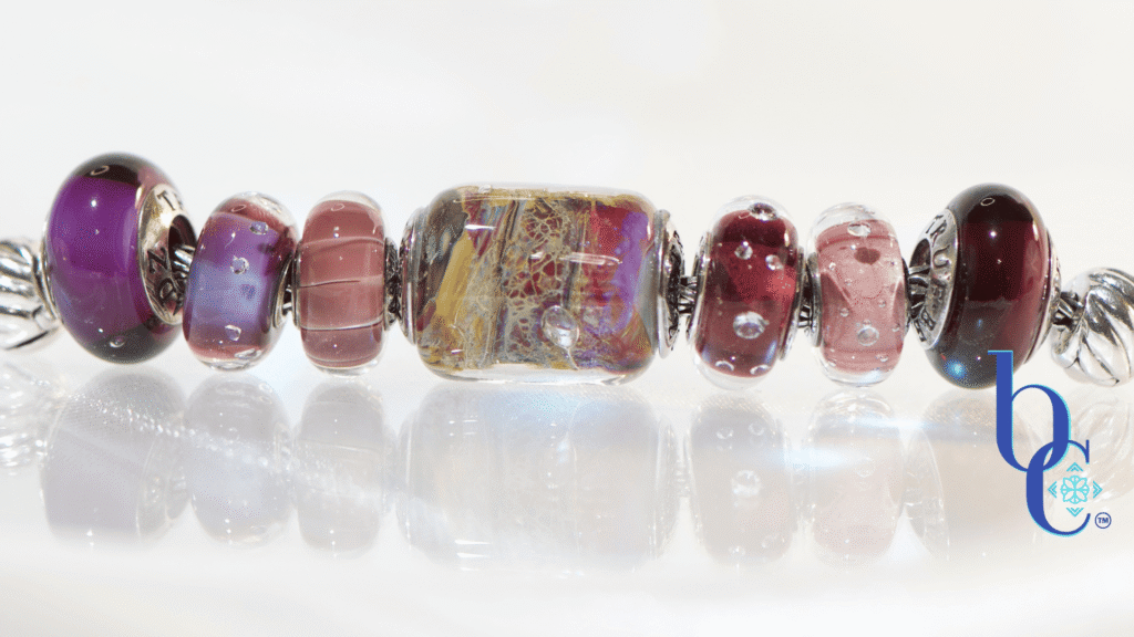 A horizontal, high-key studio photograph of an artisan charm bracelet on a silver foxtail chain. A sequence of glass and silver beads is arranged with a central complex gold, red, and yellow 'Temptation' barrel bead flanked by amethyst, pink, and deep garnet-colored glass beads, including the 'Twinkle Passion' and 'Concord Grape' designs. The beads are reflected on a clean white surface with a blue Bijoux Chat watermark logo.