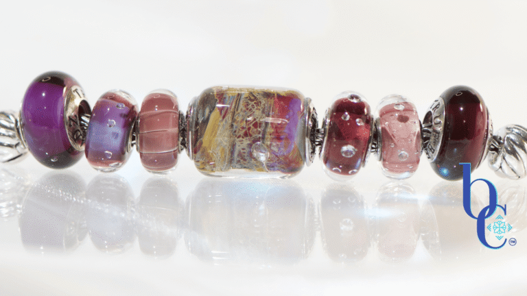 A horizontal, high-key studio photograph of an artisan charm bracelet on a silver foxtail chain. A sequence of glass and silver beads is arranged with a central complex gold, red, and yellow 'Temptation' barrel bead flanked by amethyst, pink, and deep garnet-colored glass beads, including the 'Twinkle Passion' and 'Concord Grape' designs. The beads are reflected on a clean white surface with a blue Bijoux Chat watermark logo.