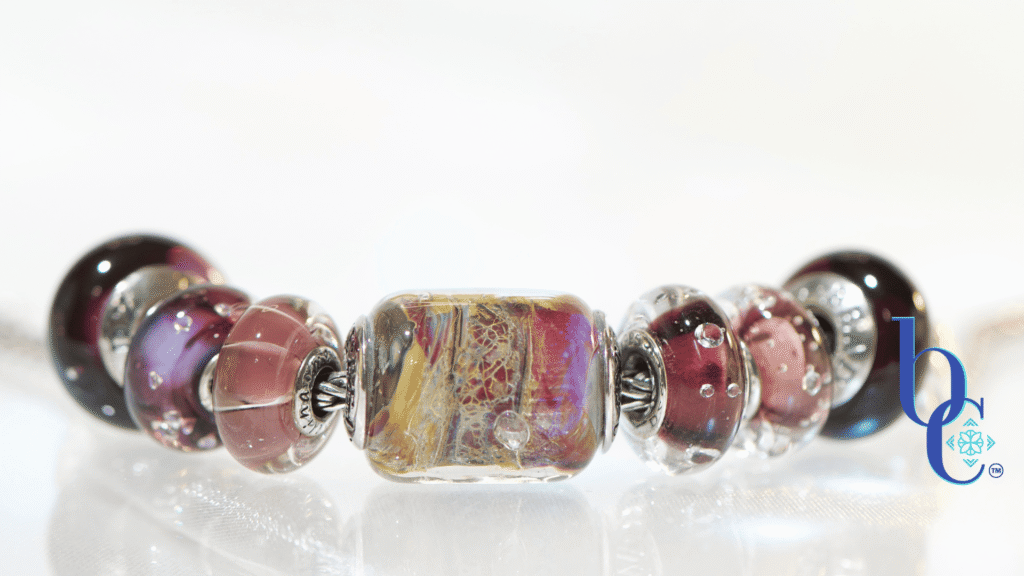 A horizontal, high-key studio photograph of an artisan charm bracelet on a silver foxtail chain. A sequence of glass and silver beads is arranged with a central complex gold, red, and yellow 'Temptation' barrel bead flanked by amethyst, pink, and deep garnet-colored glass beads, including the 'Twinkle Passion' and 'Concord Grape' designs. The beads are reflected on a clean white surface with a blue Bijoux Chat watermark logo.