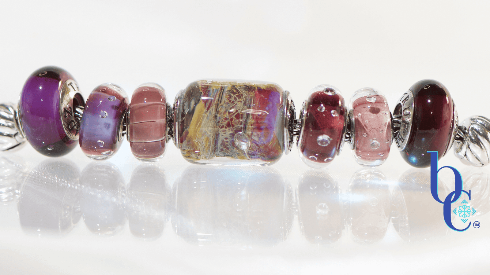 A horizontal, high-key studio photograph of an artisan charm bracelet on a silver foxtail chain. A sequence of glass and silver beads is arranged with a central complex gold, red, and yellow 'Temptation' barrel bead flanked by amethyst, pink, and deep garnet-colored glass beads, including the 'Twinkle Passion' and 'Concord Grape' designs. The beads are reflected on a clean white surface with a blue Bijoux Chat watermark logo.