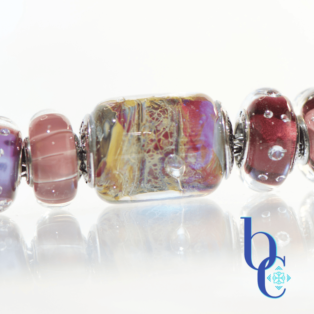 Macro photograph of a large, intricate TrueBeadz Temptation barrel glass bead with swirling gold, pink, and purple patterns, flanked by mauve and burgundy beads on a reflective surface with a BC logo.