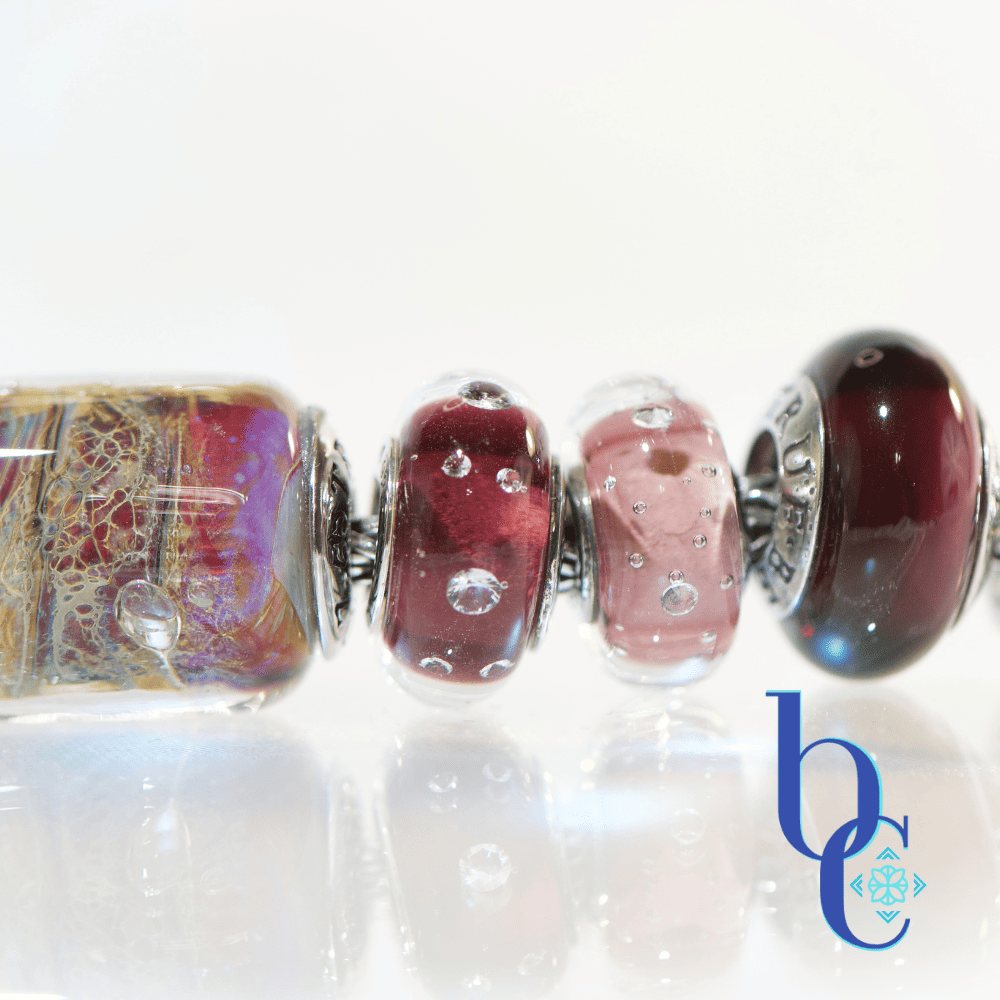 A macro photograph of the artisan glass charm bracelet detailing the deep burgundy Elfbeads Eggplant Monroe, the translucent blush Melina Kythira, and the rich cabernet TrueBeadz Amethyst on a silver chain against a reflective white background.