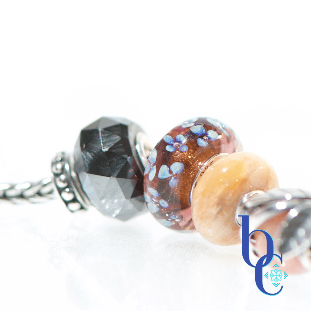 Macro studio photograph of a European charm bracelet resting on a bright, reflective white surface. The bead stack transitions from warm autumn tones on the left—featuring earthy brown glass threads, amber florals, faceted smoky quartz, and banded terracotta glass—into a central textured silver leaf charm holding a round sunstone. The right side cools down with a smooth golden quartz, blue floral glass, and a dark faceted pyrite bead. This macro features the charcoal faceted pyrite and the warm browns of the Elfbeads galaxy allure bead.