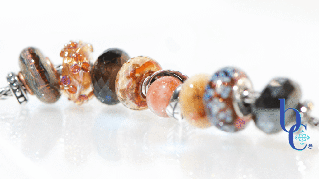 Macro studio photograph of a European charm bracelet resting on a bright, reflective white surface. The bead stack transitions from warm autumn tones on the left—featuring earthy brown glass threads, amber florals, faceted smoky quartz, and banded terracotta glass—into a central textured silver leaf charm holding a round sunstone. The right side cools down with a smooth golden quartz, blue floral glass, and a dark faceted pyrite bead.