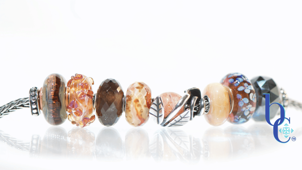 Macro studio photograph of a European charm bracelet resting on a bright, reflective white surface. The bead stack transitions from warm autumn tones on the left—featuring earthy brown glass threads, amber florals, faceted smoky quartz, and banded terracotta glass—into a central textured silver leaf charm holding a round sunstone. The right side cools down with a smooth golden quartz, blue floral glass, and a dark faceted pyrite bead.