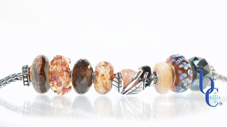Macro studio photograph of a European charm bracelet resting on a bright, reflective white surface. The bead stack transitions from warm autumn tones on the left—featuring earthy brown glass threads, amber florals, faceted smoky quartz, and banded terracotta glass—into a central textured silver leaf charm holding a round sunstone. The right side cools down with a smooth golden quartz, blue floral glass, and a dark faceted pyrite bead.