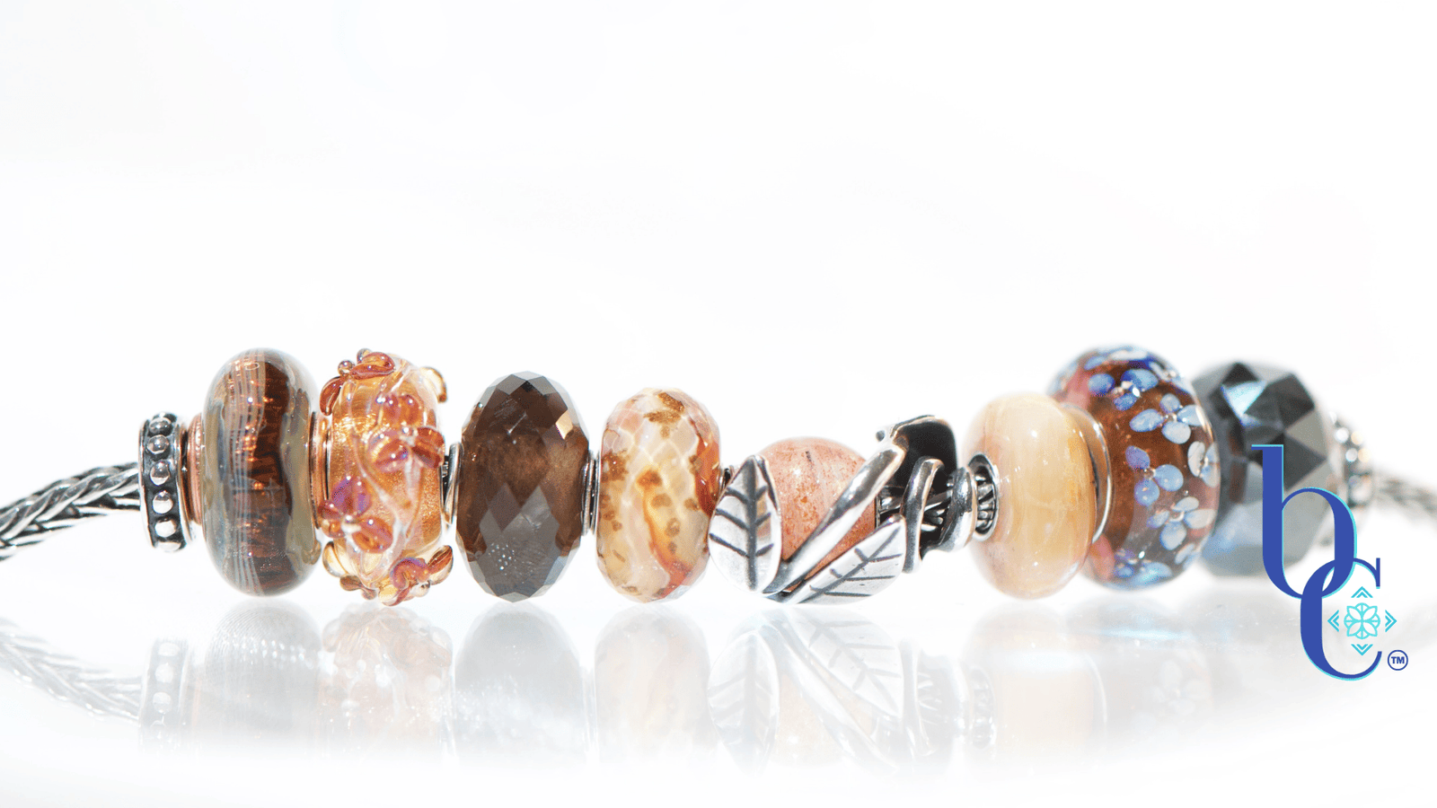 Macro studio photograph of a European charm bracelet resting on a bright, reflective white surface. The bead stack transitions from warm autumn tones on the left—featuring earthy brown glass threads, amber florals, faceted smoky quartz, and banded terracotta glass—into a central textured silver leaf charm holding a round sunstone. The right side cools down with a smooth golden quartz, blue floral glass, and a dark faceted pyrite bead.