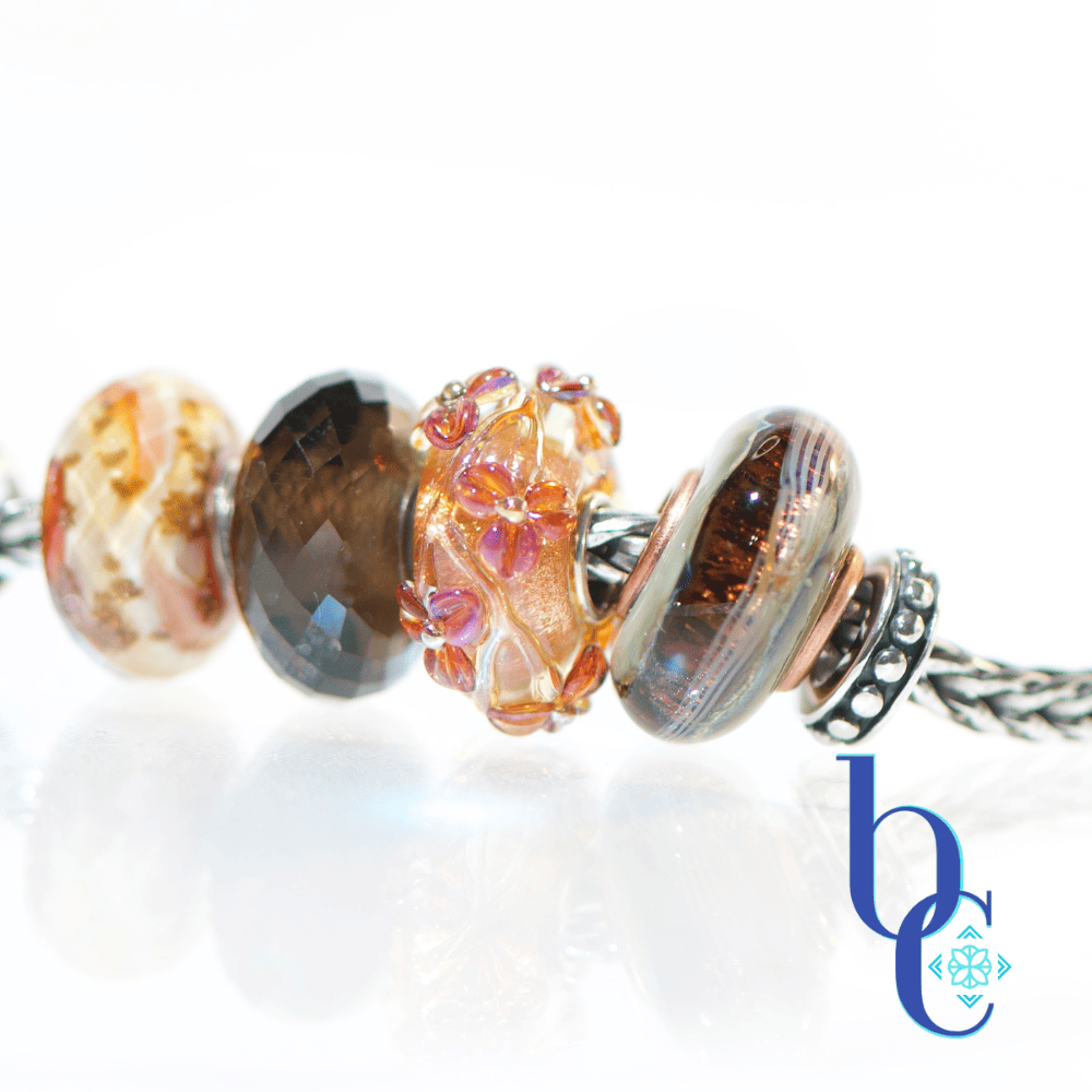 Macro studio photograph of a European charm bracelet resting on a bright, reflective white surface. The bead stack transitions from warm autumn tones on the left—featuring earthy brown glass threads, amber florals, faceted smoky quartz, and banded terracotta glass—into a central textured silver leaf charm holding a round sunstone. The right side cools down with a smooth golden quartz, blue floral glass, and a dark faceted pyrite bead. This macro features the terracotta and amber toned foral glass bead.