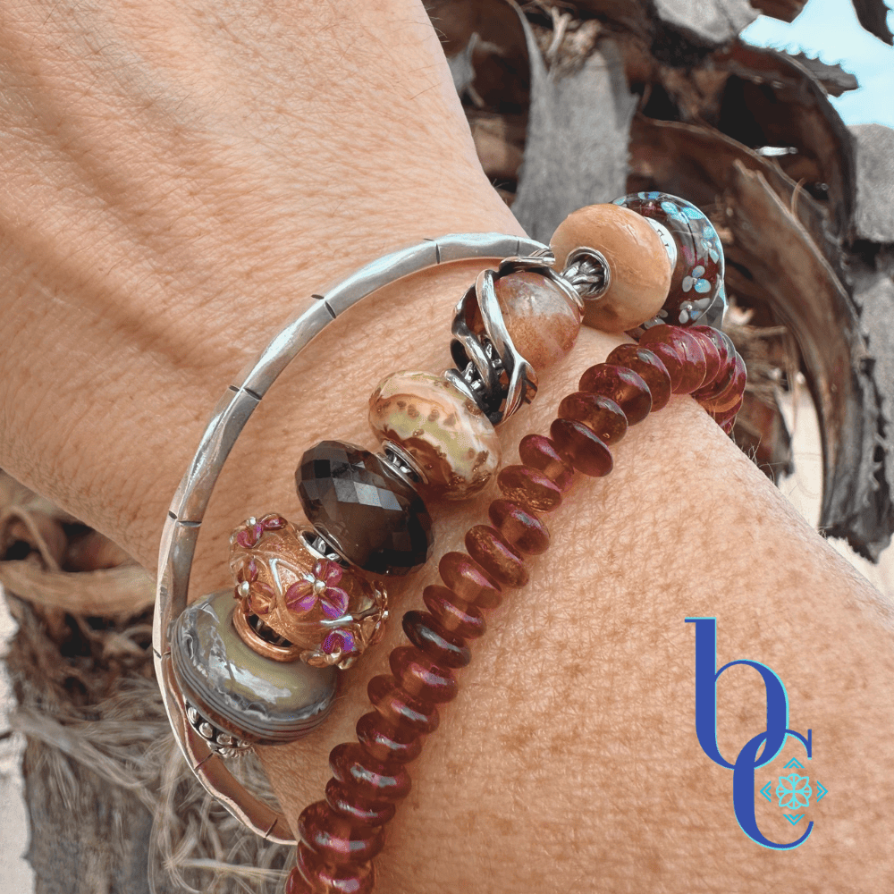 Lifestyle photograph of a wrist wearing a silver European charm bangle stacked with a natural cognac amber nugget bracelet. The charm stack features a blue and grey artisan bead, a copper-cored floral amber bead, faceted smoky quartz, and banded terracotta glass, set against an out-of-focus background of rough, natural tree bark.