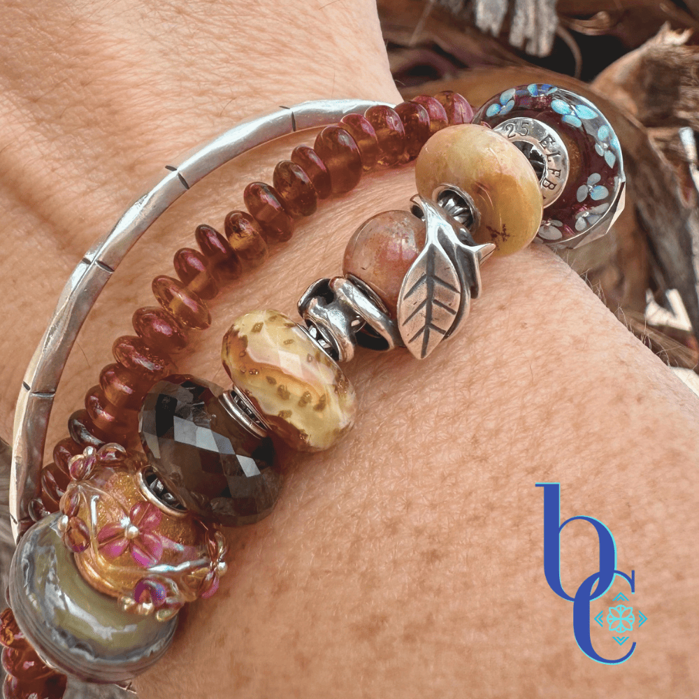 Close-up lifestyle photograph of a silver charm bracelet on a wrist, focusing on a silver leaf frame holding a round sunstone, flanked by golden quartz and dark blue floral glass beads. The silver bangle is stacked alongside a textured cognac amber bead bracelet with a natural, textured wood background.