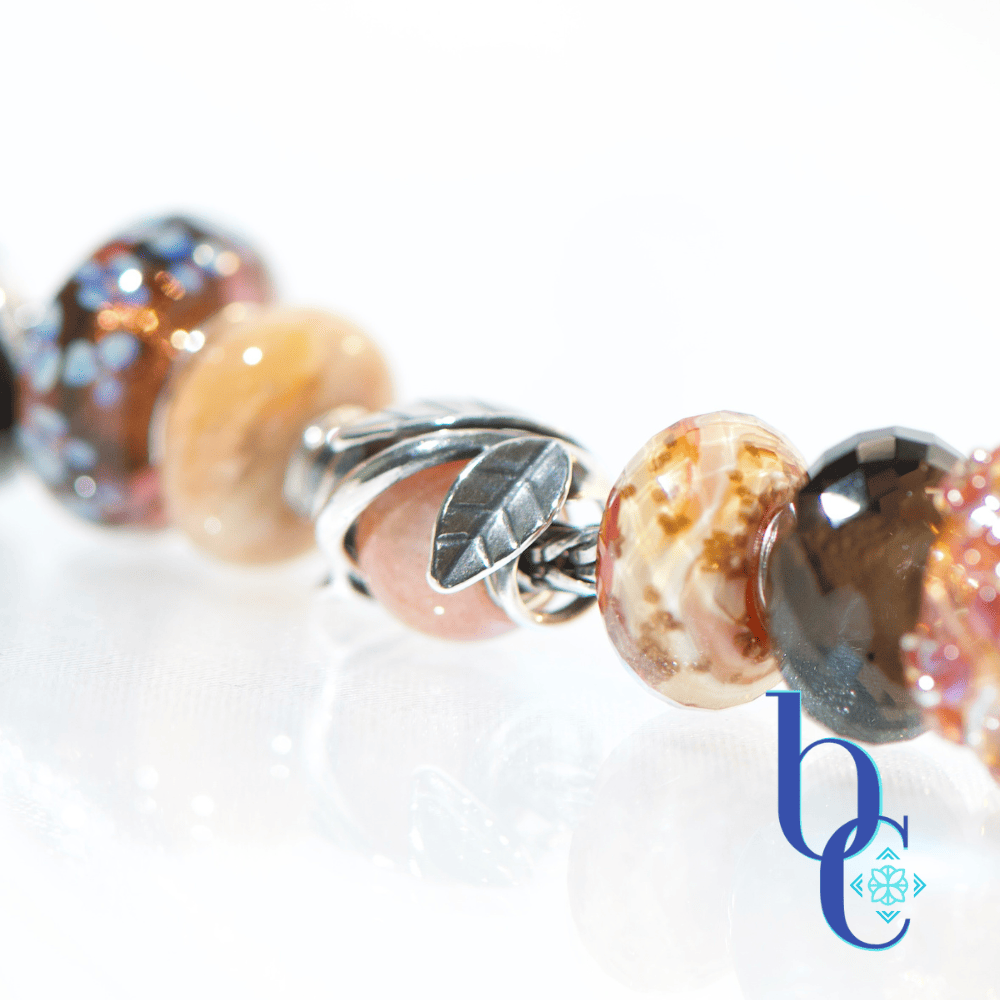 Macro studio photograph of a European charm bracelet resting on a bright, reflective white surface. The bead stack transitions from warm autumn tones on the left—featuring earthy brown glass threads, amber florals, faceted smoky quartz, and banded terracotta glass—into a central textured silver leaf charm holding a round sunstone. The right side cools down with a smooth golden quartz, blue floral glass, and a dark faceted pyrite bead. This macro features the center hero focal sunstone.