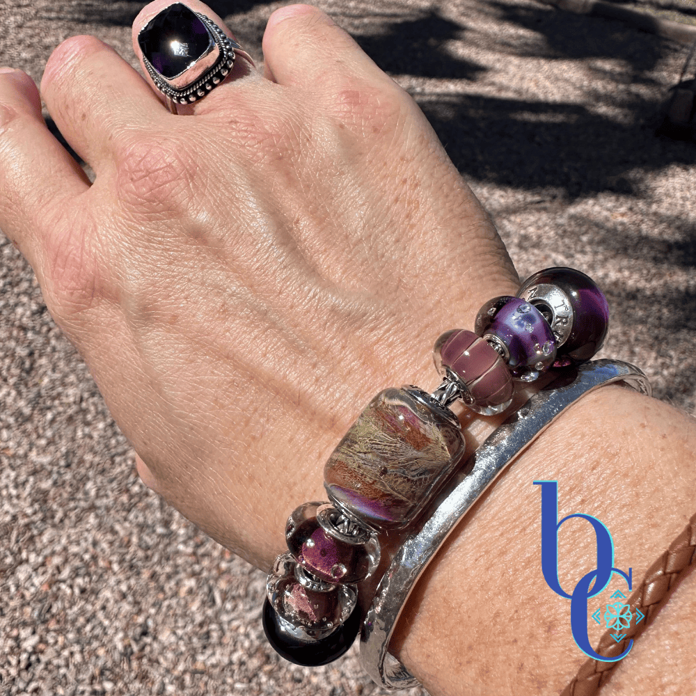 A close-up outdoor lifestyle photograph of a hand and wrist wearing the Gilded Tannin artisan glass charm bracelet, stacked with a silver hammered bangle and a braided brown leather bracelet, against a sunny gravel background with a blue BC watermark