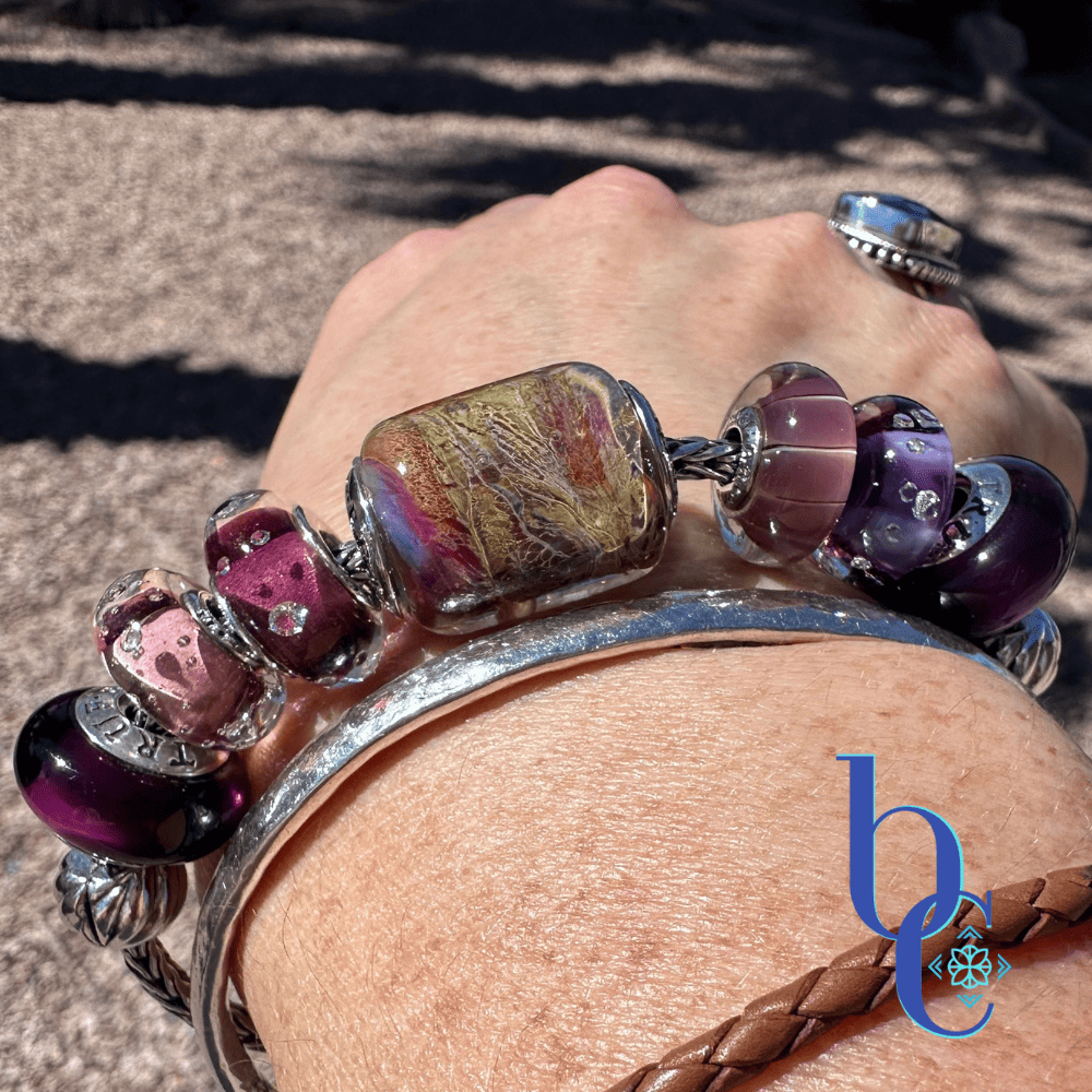 A close-up outdoor lifestyle photograph of a hand and wrist wearing the Gilded Tannin artisan glass charm bracelet, stacked with a silver hammered bangle and a braided brown leather bracelet, against a sunny gravel background with a blue BC watermark