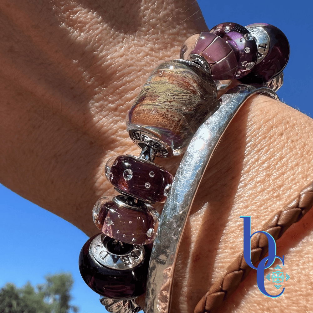 A close-up outdoor lifestyle photograph of a hand and wrist wearing the Gilded Tannin artisan glass charm bracelet, stacked with a silver hammered bangle and a braided brown leather bracelet, against a sunny gravel background with a blue BC watermark